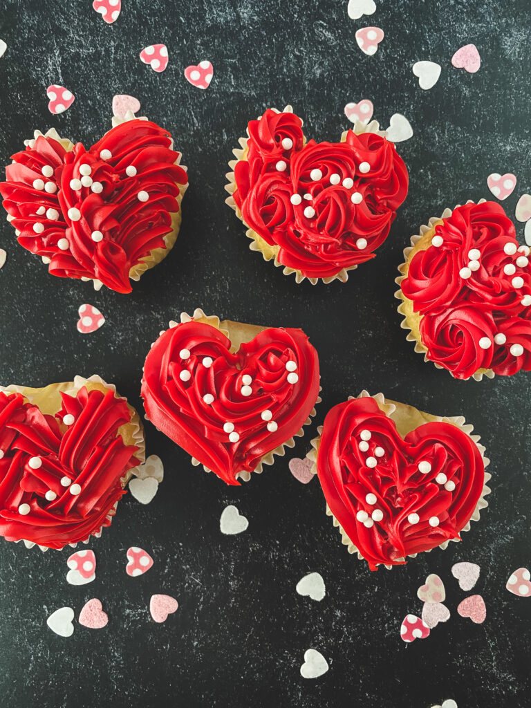 Looking for a festive edible present for Valentine’s Day? Here are our Heart-shaped Cupcakes recipes that I am certain you will adore so much. Make your loved ones feel special and full with these sweet treats.   All About Heart-shaped Cupcakes! The month of February is said to be the “Love Month”. Thus, people are busy enough looking for ways on how to make their family, friends, partners, and other significant others feel special.   Well, if you are in that scenario at this very moment, consider having edible presents for you will never go wrong with it. These heart-shaped cupcakes are perfect! They are festive in the color of red, appealing in looks, sweet in taste, and very easy to make. Even kids can join you in the kitchen making them.   All you have to do is to wrap or pack them in cute packaging and viola! You just have your beautiful cupcakes for Valentine’s day.  What Equipment Should You Prepare?  You will need the following tools in making the cupcakes:  Cupcake tin, as the mold of the cupcakes.  Cupcake Liners, to contain the batter in place.  Marbles, to be placed in the center top of the cupcake to create the curve for the shape of a heart.  Aluminum Foil, to be placed on the sides to form the heart shape.  What are the Ingredients You’ll Need?  You will prepare the following components before starting the process:  Boxed Cake Mix. Just follow the label’s instructions for the batter.  Eggs. Three pieces Oil. Set aside ⅓ cup Water. One cup Frosting Sprinkles  How to Make the Heart-shaped Cupcakes? The steps of this recipe are pretty basic and simple. Begin by, lining the cupcake tin with cupcake liners and placing a marble in the center of the "top" of the cupcake. Also, fold a small piece of foil to fit on both sides of the cupcake liner. This will create the heart mold in the baking tins.   Next, use a 1/4 measuring cup to add the batter to the cupcake tin. Bake for 15 to 18 minutes. After the time, remove and let cool in the pan for 20 to 25 minutes.   Once removed you should have a heart-shaped cupcake! Frost as you would like and well done! You just made your own heart-shaped cupcakes for Valentine’s.  How to Decorate the Cupcakes? In this recipe, there are three decorating tips that I can offer. First, to make the mini rosettes, hold your piping bag straight up and down and make small circles using a "star tip". Second, for the Criss/Cross Design, slightly angle the piping bag and create a criss-cross pattern. And ultimately, for the heart outline, outline the cupcake and make small circles in the center to fill in the space. You can also add sprinkles!